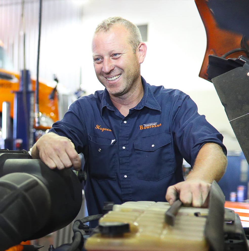 Un mécanicien souriant en uniforme dans un garage, entouré de cabines de camions Transport Bourassa et d'équipement de maintenance de flotte.