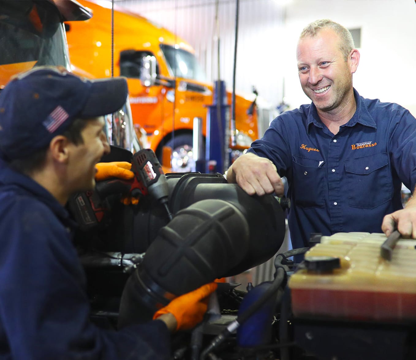 Un mécanicien souriant en uniforme dans un garage, entouré de cabines de camions Transport Bourassa et d'équipement de maintenance de flotte.